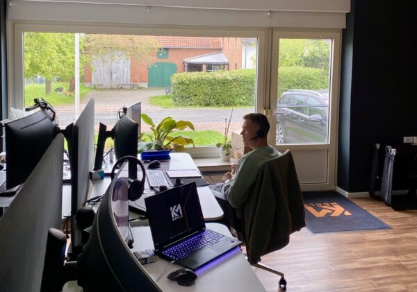 Mitarbeiter mit Headset sitzt in einem modernen Büro mit mehreren Arbeitsplätzen und Curved-Monitoren, vor einem großen Fenster mit Blick auf eine ruhige Wohnstraße.