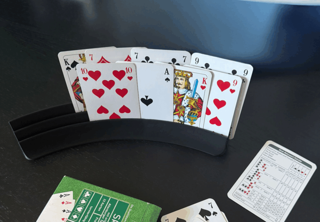 Ein Skat-Spiel ist im Gange: Auf einem dunklen Holztisch stehen Spielkarten in einem schwarzen Kartenhalter. Im Vordergrund liegen eine grüne Skat-Kartenverpackung, einige abgelegte Karten sowie ein Skatblock mit Punkten. Im Hintergrund befinden sich weitere Alltagsgegenstände.